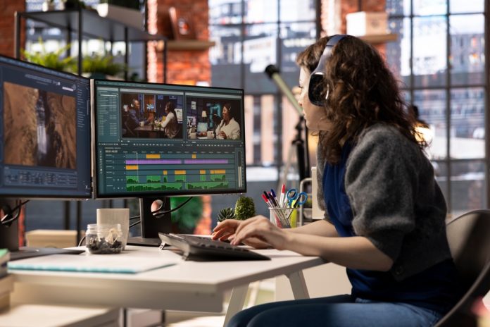 a woman sitting at a desk with two monitors, editing a video
